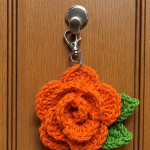 Peut inclure: Une fleur de rose orange crochet&eacute;e avec deux feuilles vertes attach&eacute;e &agrave; un porte-cl&eacute;s &agrave; fermoir mousqueton en m&eacute;tal argent&eacute;. La fleur pr&eacute;sente des p&eacute;tales superpos&eacute;s cr&eacute;&eacute;s avec du fil, et la quincaillerie m&eacute;tallique inclut un anneau.