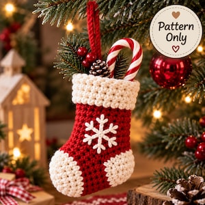 Puede incluir: Un adorno navideño de calcetín tejido a ganchillo en rojo y blanco con un motivo de copo de nieve blanco, puño blanco y lazo rojo para colgar. El calcetín contiene un bastón de caramelo, una piña pequeña y bayas rojas. Una etiqueta circular dice Pattern Only.