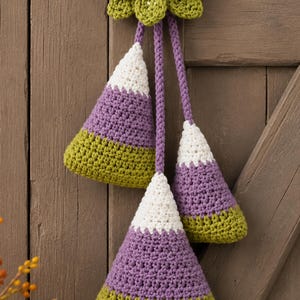 Pode incluir: Uma decora&ccedil;&atilde;o de croch&ecirc; suspensa com tr&ecirc;s ornamentos triangulares em forma de milho doce nas cores branco, roxo e verde-oliva. Os ornamentos est&atilde;o suspensos por cord&otilde;es tran&ccedil;ados roxos a partir de um la&ccedil;o central roxo e folhas de croch&ecirc; verdes.
