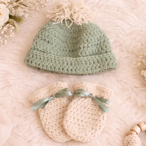 Puede incluir: Conjunto de bebé tejido a crochet en colores suaves. El conjunto incluye un gorro verde claro con un pompón color crema y dos mitones color crema atados con cintas verde claro. Los artículos se muestran sobre una superficie blanca y esponjosa.