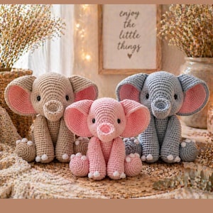 May include: Three crocheted elephant stuffed animals in beige, pink, and gray, each with large, contrasting pink ears. The elephants are sitting on a woven mat, with a framed sign in the background that reads "enjoy the little things."