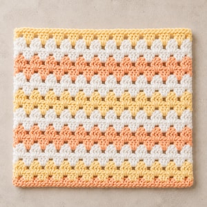 May include: A square crochet piece featuring a repeating granny stitch pattern. The design consists of horizontal stripes in alternating colors of soft yellow, crisp white, and light peach, creating a textured and uniform geometric textile surface.