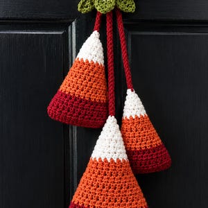 Pode incluir: Uma decora&ccedil;&atilde;o de croch&ecirc; suspensa com tr&ecirc;s ornamentos em forma de doce de milho nas cores branca, laranja e vermelho escuro. Os ornamentos est&atilde;o suspensos por cord&otilde;es de l&atilde; vermelha a um la&ccedil;o central bege e folhas verdes de croch&ecirc;.