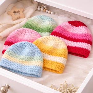 May include: A collection of colorful, crocheted beanies in a white drawer. The beanies are in shades of pink, red, yellow, green, and blue. The hats are arranged on a white fabric. A wooden star and decorative beads are also in the drawer.