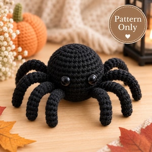 Pode incluir: Uma aranha de crochê preta com dois olhos pretos grandes e brilhantes. A aranha tem um corpo redondo e oito pernas curvas. Um emblema circular no canto superior direito contém o texto Pattern Only com um pequeno ícone de coração abaixo.