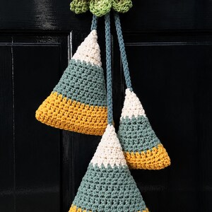 Pode incluir: Um enfeite de parede em croch&ecirc; com um la&ccedil;o rosa e folhas verdes no topo, com tr&ecirc;s ornamentos triangulares pendentes. Cada pe&ccedil;a apresenta blocos de cores em branco, verde s&aacute;lvia e amarelo mostarda, suspensos por cord&otilde;es de fio tran&ccedil;ado.