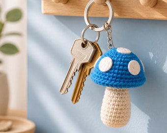 Peut inclure: Un porte-clés champignon en crochet avec un chapeau bleu vif à pois blancs et un pied beige. Le champignon est fixé à un anneau en métal argenté retenant deux clés métalliques standard, suspendu à une patère murale en bois.