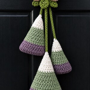 Pode incluir: Uma decora&ccedil;&atilde;o de parede em croch&ecirc; suspensa com tr&ecirc;s ornamentos em forma de cone nas cores branco, verde s&aacute;lvia e roxo. Os cones est&atilde;o suspensos por cord&otilde;es de croch&ecirc; verdes a partir de um topo central bege em forma de la&ccedil;o.