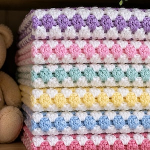 May include: A stack of six folded crochet blankets featuring a repeating pattern of white yarn and alternating pastel-colored yarn rows in purple, pink, mint green, yellow, light blue, and a darker pink, all showcasing a textured bobble stitch design.