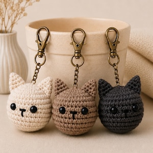 May include: Three crochet cat head keychains in cream, brown, and black. Each cat has black button eyes and a stitched nose and mouth. The keychains have bronze-colored clasps and chains.