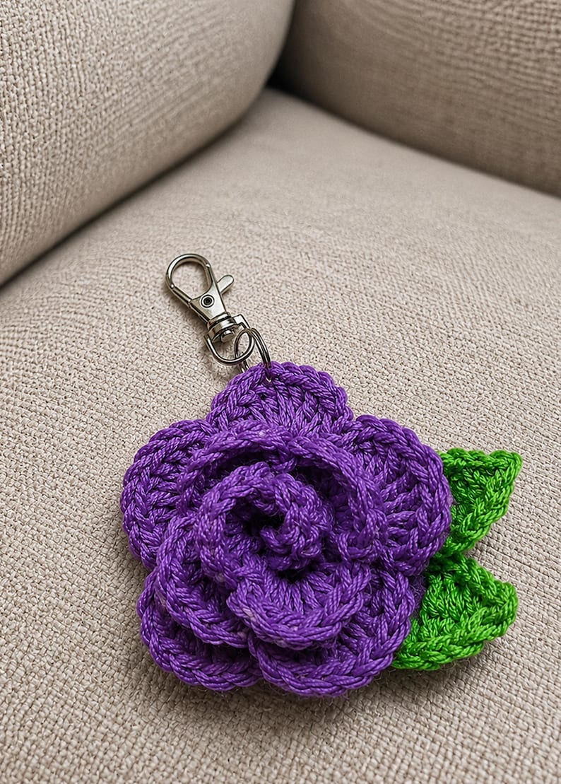 Peut inclure: Un porte-cl&eacute;s fleur crochet&eacute; violet pr&eacute;sentant un motif de rose en couches avec deux feuilles crochet&eacute;es vert vif attach&eacute;es sur le c&ocirc;t&eacute; et un fermoir mousqueton en m&eacute;tal argent&eacute; fix&eacute; au sommet de la pi&egrave;ce florale.