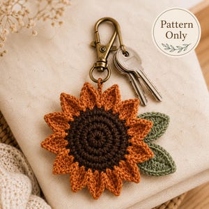 May include: A crochet sunflower keychain featuring a dark brown center, orange petals, and two green leaves. The sunflower is attached to a bronze-colored metal lobster clasp with two silver keys. A circular label displays the text Pattern Only.