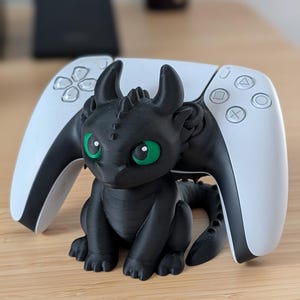 Peut inclure: Une figurine de dragon noir aux yeux verts est placée devant une manette blanche de PlayStation 5. Le dragon a une surface texturée et détaillée. La manette a des boutons noirs et une finition lisse. Les objets sont sur une surface en bois.