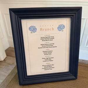 May include: A navy blue framed menu for an Easter brunch. The menu lists starters, mains, sides, desserts, and drinks. The menu includes items like Spring Vegetable Soup, Baked Salmon, and Easter Egg Cupcakes. The frame is angled.