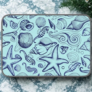 May include: A rectangular tin box featuring a light blue background with a repeating dark blue pattern of various seashells, starfish, and seahorses. The design includes detailed line work on each marine creature and shell across the surface.