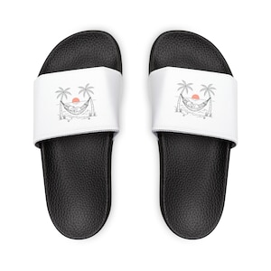 May include: Black slide sandals with a white strap featuring a black and white illustration of a hammock between palm trees, with a sun in the background. The sandals have a textured footbed.