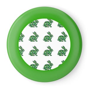 May include: A green frisbee with a white center featuring a repeating pattern of green dotted rabbits. The frisbee has a solid green rim and a smooth, flat surface. The rabbits are in a hopping pose.