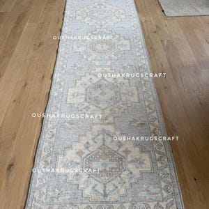 Peut inclure: Un tapis de couloir rectangulaire gris clair avec un motif géométrique. Le tapis présente un motif central avec des accents beiges, bleus et rouille. Le texte "OUSHAKRUGCRAFT" est imprimé sur le tapis.