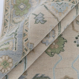 Op de afbeelding: Close-up van een opgevouwen beige tapijt met een gedetailleerd bloemenpatroon. Het tapijt heeft een lichtblauwe rand en accenten, met groene en bruine bloemmotieven. De textuur van het tapijt is zichtbaar, wat wijst op een zacht, geweven materiaal.