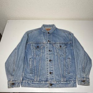 May include: A light blue denim jacket featuring a classic collar, a front button closure, two chest pockets with buttoned flaps, and two vertical side pockets. The jacket has long sleeves with buttoned cuffs and a small red brand tab on the chest.