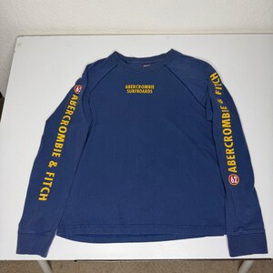 May include: A long-sleeve navy blue crewneck shirt featuring yellow text that reads ABERCROMBIE SURFBOARDS on the chest. Both sleeves display the text ABERCROMBIE & FITCH in yellow, accompanied by a small circular red and white logo with the number 62.