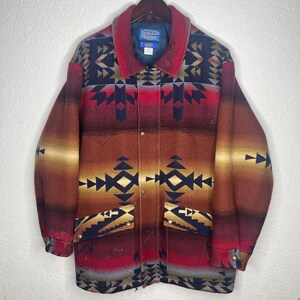 May include: A long-sleeve button-up jacket featuring a multi-colored geometric pattern in shades of red, brown, black, and cream. The jacket includes a pointed collar, two front pockets with button closures, and a blue interior brand label.