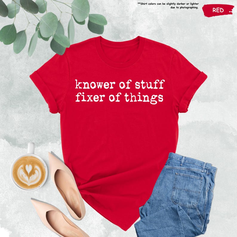 May include: A red short-sleeved t-shirt with white text printed on the front that reads "knower of stuff fixer of things". The shirt features a crew neckline and is displayed alongside a cup of coffee and blue denim jeans.