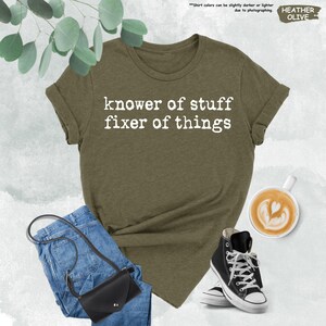 May include: A heather olive t-shirt with white text that reads "knower of stuff fixer of things" in a typewriter font. The shirt is paired with blue denim jeans, a black crossbody bag, and black and white high-top canvas sneakers.