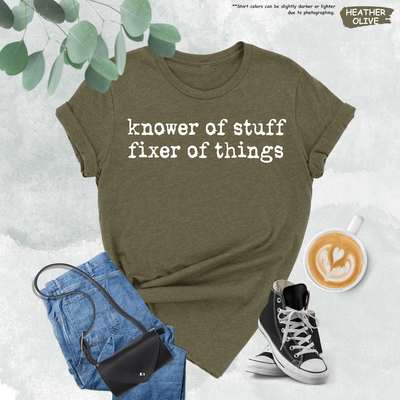 May include: A heather olive t-shirt with white text that reads "knower of stuff fixer of things" in a typewriter font. The shirt is paired with blue denim jeans, a black crossbody bag, and black and white high-top canvas sneakers.