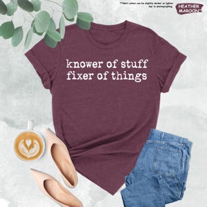 May include: A heather maroon t-shirt featuring the white text "knower of stuff fixer of things" printed on the front. The shirt is displayed alongside a cup of coffee with latte art and a pair of light-colored pointed-toe heels and blue jeans.