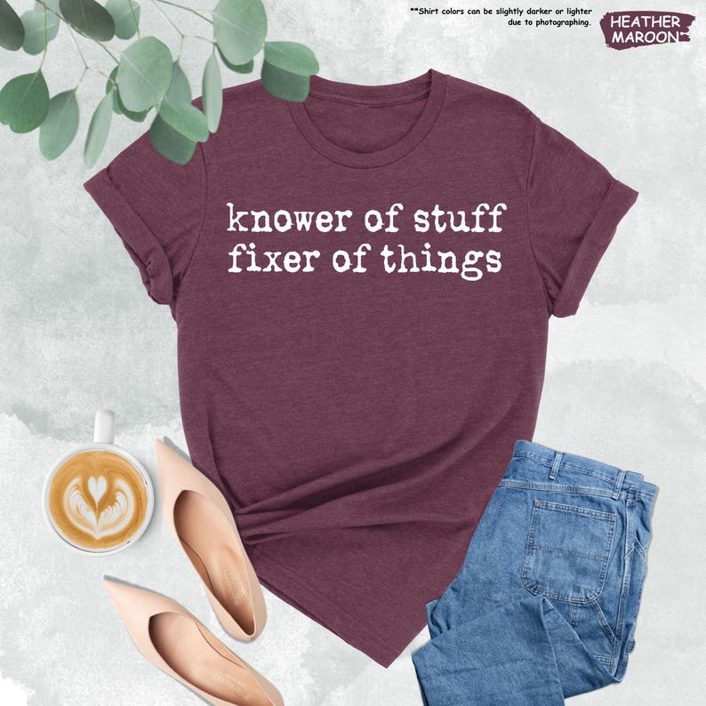 May include: A heather maroon t-shirt featuring the white text "knower of stuff fixer of things" printed on the front. The shirt is displayed alongside a cup of coffee with latte art and a pair of light-colored pointed-toe heels and blue jeans.