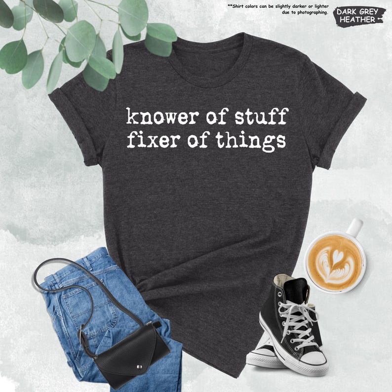 May include: A dark grey heather t-shirt with white text that reads "knower of stuff fixer of things" in a typewriter-style font. The shirt is displayed alongside blue denim jeans, a black crossbody bag, and a white mug of coffee.
