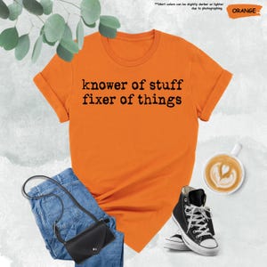 May include: An orange short-sleeved t-shirt with the black text "knower of stuff fixer of things" printed on the front. The shirt is displayed alongside blue denim jeans, a black crossbody bag, and a white mug filled with coffee.