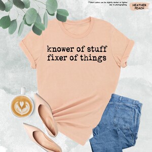 May include: A heather peach short-sleeve t-shirt with the black text "knower of stuff fixer of things" printed on the front. The shirt features a crew neckline and is displayed alongside a cup of coffee and a pair of blue denim jeans.
