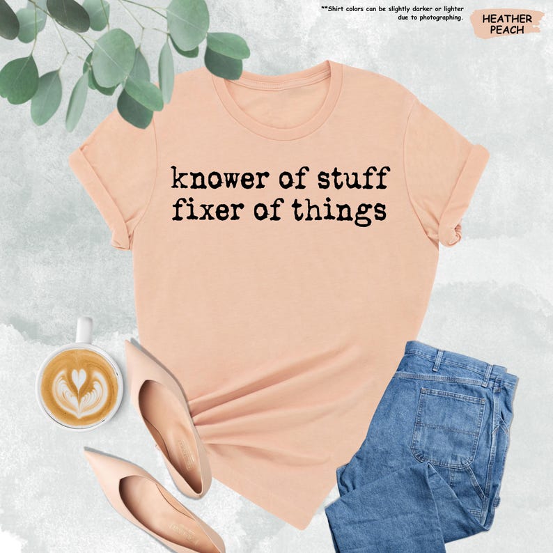 May include: A heather peach short-sleeve t-shirt with the black text "knower of stuff fixer of things" printed on the front. The shirt features a crew neckline and is displayed alongside a cup of coffee and a pair of blue denim jeans.