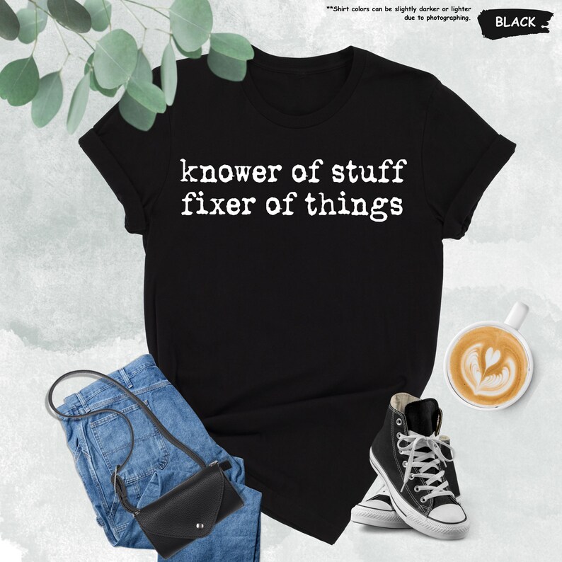 May include: A black short-sleeve t-shirt with white text printed on the front that reads "knower of stuff fixer of things". The shirt is displayed alongside blue denim jeans, a black crossbody bag, and a pair of black and white high-top sneakers.