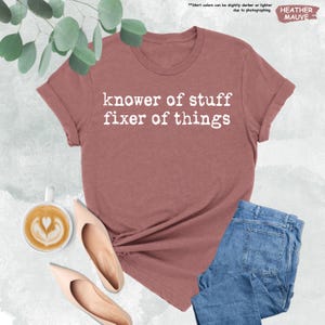 May include: A heather mauve short-sleeve t-shirt with the white text "knower of stuff fixer of things" printed on the front. The shirt is displayed alongside a cup of coffee with latte art, two beige pointed-toe heels, and blue denim jeans.