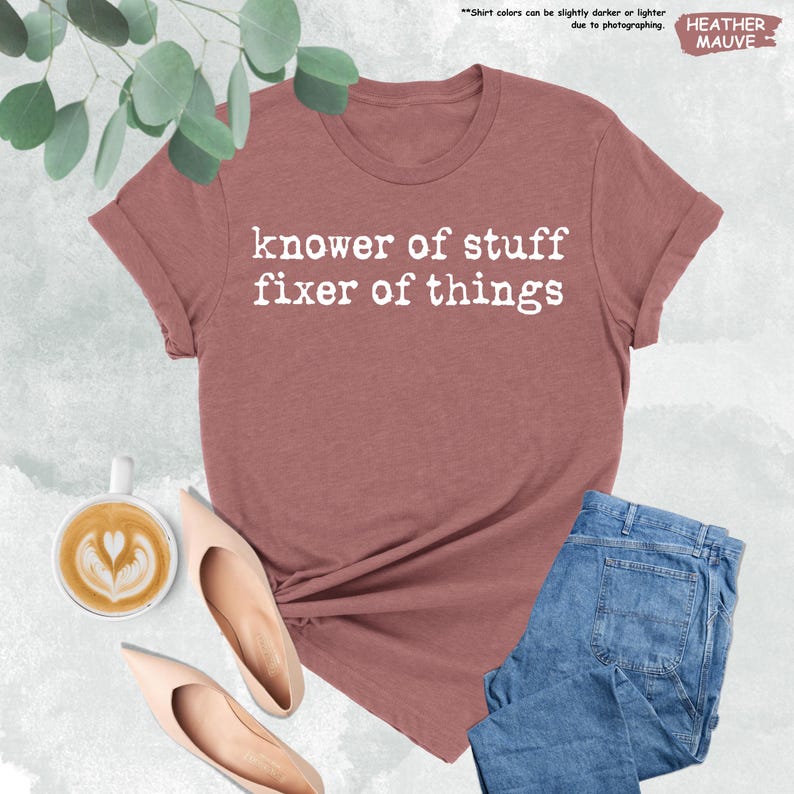 May include: A heather mauve short-sleeve t-shirt with the white text "knower of stuff fixer of things" printed on the front. The shirt is displayed alongside a cup of coffee with latte art, two beige pointed-toe heels, and blue denim jeans.
