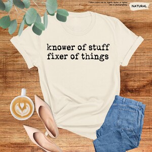 May include: A natural-colored short-sleeve t-shirt featuring the black text "knower of stuff fixer of things" printed in a typewriter-style font across the chest. The shirt is displayed alongside a cup of coffee and blue denim jeans.
