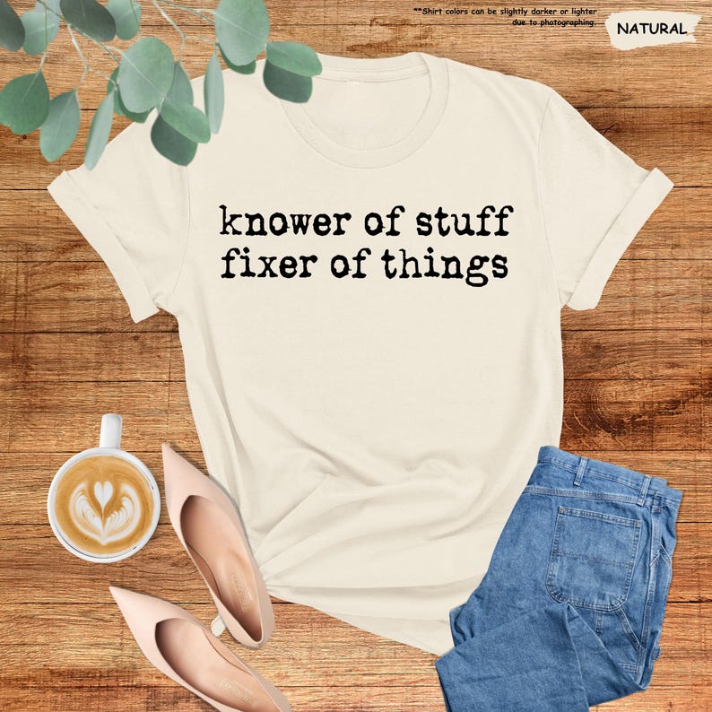 May include: A natural-colored short-sleeve t-shirt featuring the black text "knower of stuff fixer of things" printed in a typewriter-style font across the chest. The shirt is displayed alongside a cup of coffee and blue denim jeans.