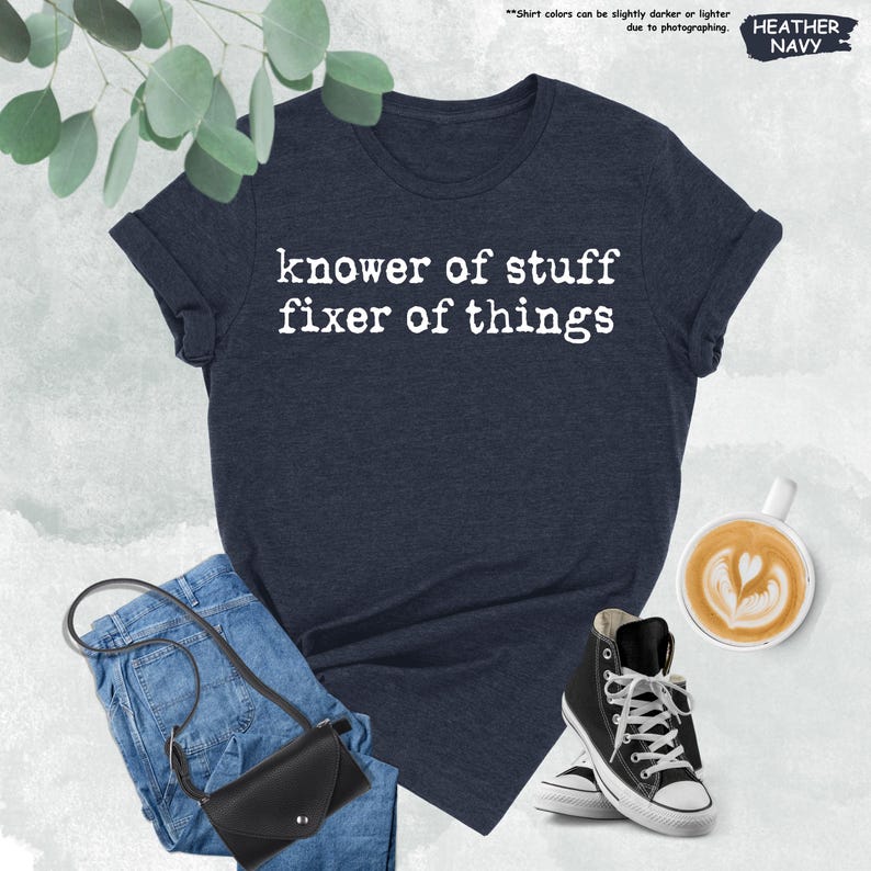 May include: A heather navy blue short-sleeve t-shirt with the white text "knower of stuff fixer of things" printed on the front. The shirt is displayed alongside blue denim jeans, a black crossbody bag, and a cup of coffee with latte art.