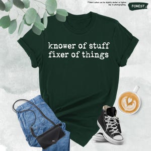 May include: A forest green short-sleeved t-shirt with the white text "knower of stuff fixer of things" printed on the chest. The shirt is displayed alongside blue denim jeans, a black crossbody bag, and a cup of coffee with latte art.