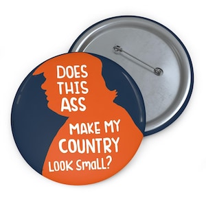 May include: A round button with a navy blue background and an orange silhouette of a person. The text "DOES THIS ASS MAKE MY COUNTRY LOOK SMALL?" is in white. The back of the button is silver with a pin.