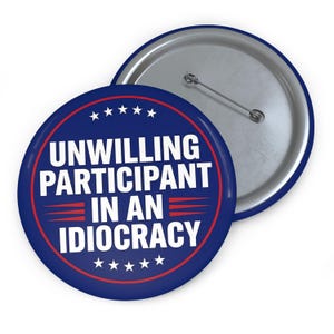 May include: A round, blue button with the text "UNWILLING PARTICIPANT IN AN IDIOCRACY" in white. The text is surrounded by a red border and stars. The button has a silver pin back.
