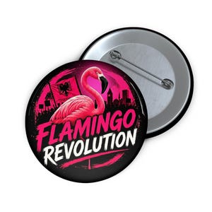 May include: A round pin-back button featuring a vibrant pink flamingo graphic against a dark background. The design includes a silhouette of a city skyline, a flag with a double-headed eagle, and the bold white and pink text FLAMINGO REVOLUTION.
