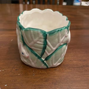 May include: A small, white ceramic planter with a scalloped rim. The planter is decorated with green leaves and a small bunny face. The interior is white. A decorative item for plants or small items.