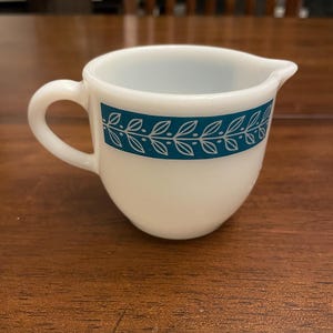 May include: A white creamer with a handle and a spout. A teal band with a leaf design encircles the creamer. The creamer is sitting on a wooden surface.