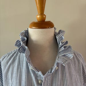May include: A blue and white striped shirt with a ruffled collar is displayed on a mannequin. The shirt has a button-down front and a classic, preppy style. The mannequin's neck is covered in a white fabric.