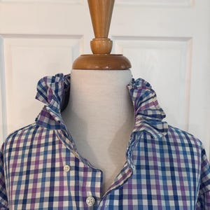 May include: A long-sleeved button-down shirt featuring a blue, purple, and white gingham check pattern. The shirt is designed with a high, ruffled collar and includes two visible white buttons on the front placket.