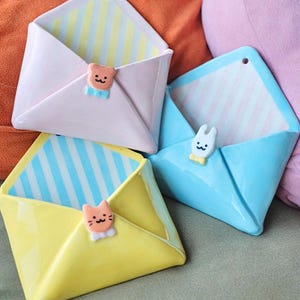 May include: Three ceramic wall-mounted envelope holders in pastel pink, light blue, and yellow. Each envelope features a striped interior and a small, sculpted animal figure—a bear, a rabbit, and a cat—attached to the centre flap closure.